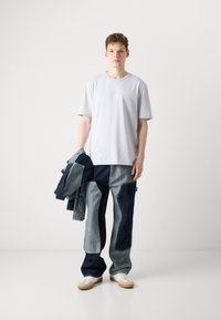 Light gray crew neck t-shirt with a small logo, paired with patchwork denim jeans in various shades of blue and a dark denim jacket.