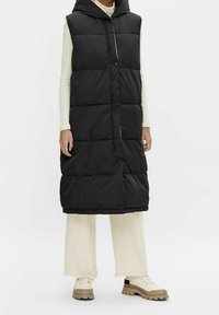 Doudoune noire sans manches avec capuche, dotée d'une fermeture à boutons et d'une texture matelassée. Portée sur un haut à manches longues crème et un pantalon ample beige.