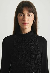 Robe noire avec un broderie florale texturée, présentant une silhouette ajustée et des manches longues, superposée sur un col roulé noir. Boucles d'oreilles simples en forme de Créole.
