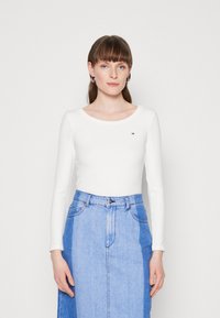 Frau mit heller Haut, braunem Haar mit Pony, trägt silberne Creolen, ein tailliertes weißes Langarmshirt und einen zweifarbigen blauen Denimrock.
