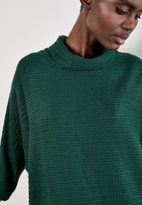 Maglione verde lavorato a maglia con collo alto, caratterizzato da un motivo testurizzato e una vestibilità ampia. Il tessuto è morbido e ha un design a costine.