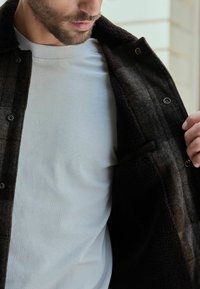 Uomo con camicia bianca solleva una giacca scura con bottoni per rivelare il rivestimento interno testurizzato e la tasca della giacca.