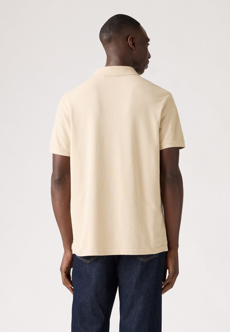 Camisa polo beige hecha de tela suave, de mangas cortas y con un cuello clásico. Combinada con jeans de mezclilla oscuros, vista desde la parte de atrás.