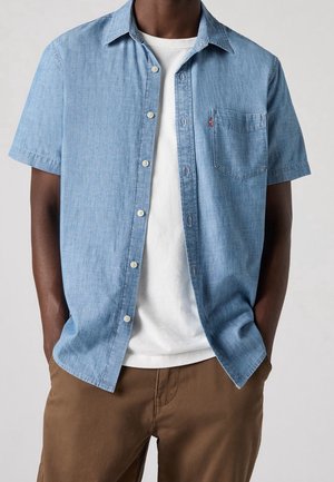 Mann trägt ein hellblaues kurzärmeliges Hemd über einem weißen T-Shirt und braune Hose, die Hände in den Taschen, vor neutralem Hintergrund.