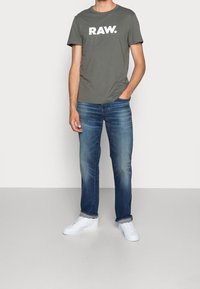 Olivgrön t-shirt med rund halsringning och vit "RAW." logotyp, kombinerad med blå denimjeans och vita sneakers. Avslappnad passform och enkelt design.