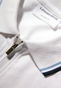 Weißes Poloshirt mit Reißverschlusskragen, verziert mit blauen und marineblauen Streifen entlang der Ränder des Kragens; strukturierter Stoff mit einem glatten Reißverschluss.