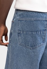 Το αριστερό χέρι ακουμπά κοντά στην πίσω τσέπη ανοιχτόχρωμου τζιν τζιν με λευκή ριγωτή μπλούζα.