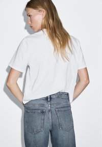 Weißes Kurzarm-T-Shirt mit lockerer Passform, kombiniert mit hoch taillierten, ausgewaschenen blauen Jeans mit doppelten Gesäßtaschen und minimaler Naht.