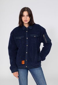 Granatowa polarowa kurtka o teksturowanej powierzchni, z zatrzaskami, dwoma kieszeniami na piersi oraz kieszenią na suwak na rękawie. Zawiera pomarańczową metkę.