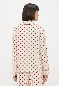 Žena s krátkými tmavými vlasy, oblečená v krémových pyžamech s červenými puntíky, zobrazená zezadu na jednolitém pozadí.