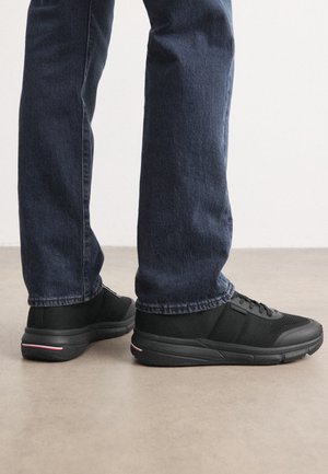 Person wearing black sneakers with textured sides and dark blue jeans standing on a smooth gray floor against a white wall.