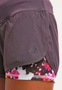 Lila idrottsshorts med en flerlagersdesign, pryds av en färgglad blommönstring undertill och en Adidas-logotyp på sidan. Slät textur.