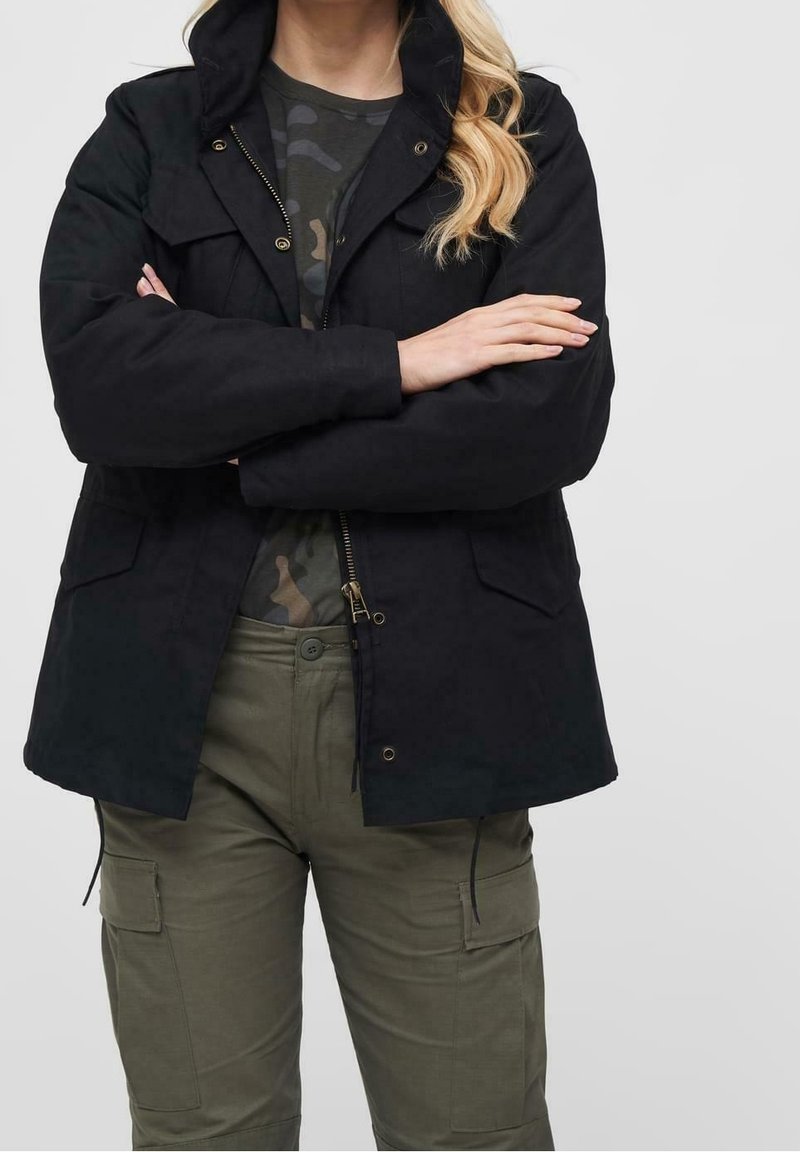 Femme vêtue d'une veste noire par-dessus un t-shirt camouflage avec un pantalon cargo vert olive, bras croisés, sur un fond uni.