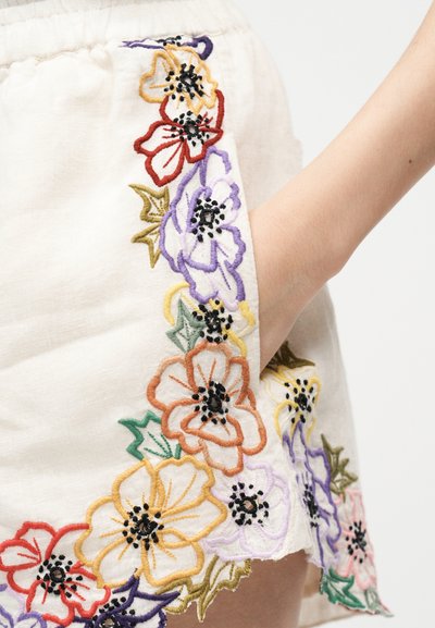 Main de femme dans la poche d'un short crème décoré de fleurs multicolores brodées sur le côté et l'ourlet.