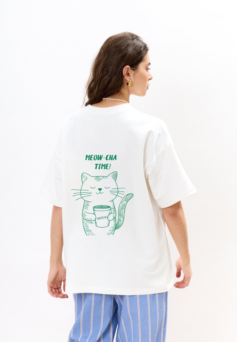 Witte katoenen t-shirt met korte mouwen, voorzien van een groene kat graphic die een beker vasthoudt en de tekst "MEOW-CHA TIME!" op de rug.