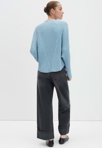 Donna in piedi di spalle, che indossa un maglione a costine blu chiaro, pantaloni neri a gamba larga con risvolto e scarpe nere slip-on.