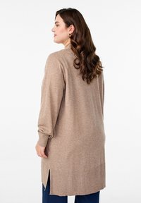 Suéter beige de punto con aberturas laterales, mangas largas y puños acanalados. Textura suave, corte ligeramente holgado. Llevado sobre pantalones azul oscuro.