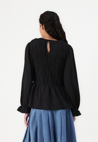 Black smocked blouse with long sleeves and a keyhole back detail, paired with a blue pleated skirt. Textured fabric with a flowy silhouette.