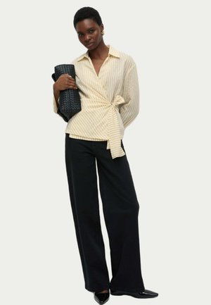 Blusa avvolgente a strisce color avorio con chiusura a legaccio, abbinata a pantaloni ampi neri e tacchi a punta neri. Gli accessori includono una borsa nera a trama.