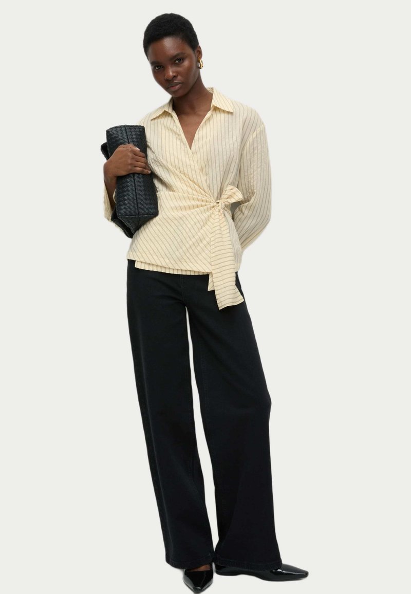Blusa avvolgente a strisce color avorio con chiusura a legaccio, abbinata a pantaloni ampi neri e tacchi a punta neri. Gli accessori includono una borsa nera a trama.