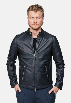 Jeune homme aux cheveux blonds courts portant une veste en cuir noire zippée et un pantalon foncé, les mains dans les poches, regardant droit devant.