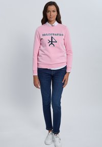 Sudadera rosa con el logo "VALCUATRO" en azul marino, cuello redondo, puños acanalados, combinada con vaqueros pitillo azul oscuro y zapatillas blancas.