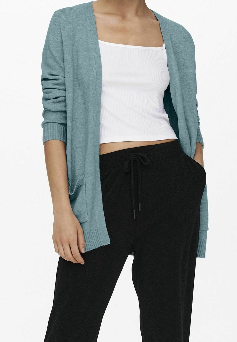 Femme portant un cardigan bleu clair, un débardeur blanc court et un pantalon noir à cordon, une main dans la poche.