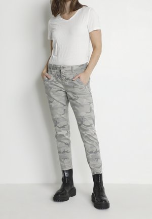 Femme portant un t-shirt blanc à manches courtes en V, un pantalon camouflage gris avec des boutons et des bottines noires épaisses, debout contre un mur blanc.