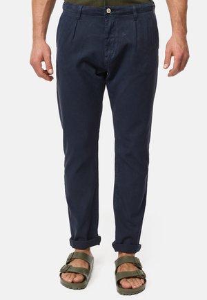 Pantaloni di cotone blu navy con frontale plissettato, gambe affusolate, orli ai polsi, abbinati a sandali verde oliva con fibbie.