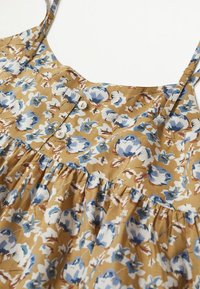 Robe sans manches à bretelles réglables, fond beige, motif floral bleu et blanc, avec deux boutons blancs sur la poitrine.