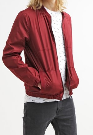 Blonde Person trägt eine rote Jacke mit Reißverschluss über einem weißen T-Shirt mit Papierflugzeugmuster und dunkelgrauen Hosen, Hände in den Taschen.