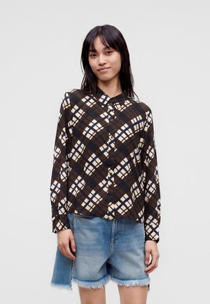 Jeune femme portant une chemise à carreaux marron boutonnée et un short en jean effiloché, debout devant un fond clair uni.