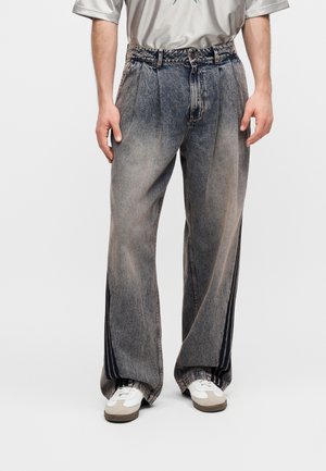 Homme portant un jean gris délavé à coupe ample avec des bandes noires sur les côtés, des baskets blanches et un t-shirt gris clair avec un logo étoile.