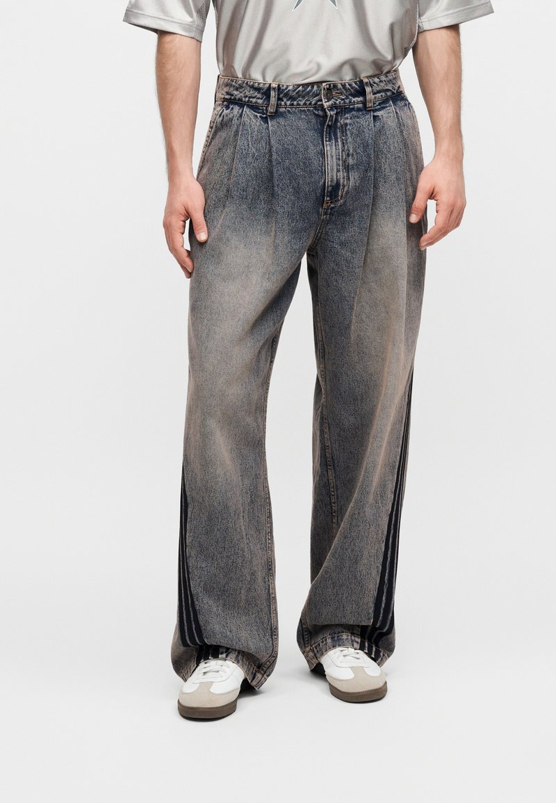 Άνδρας που φοράει άνετο ξεθωριασμένο γκρι τζιν με μαύρες λωρίδες στο πλάι, λευκά αθλητικά παπούτσια και ανοιχτό γκρι μπλουζάκι με λογότυπο αστεριού.