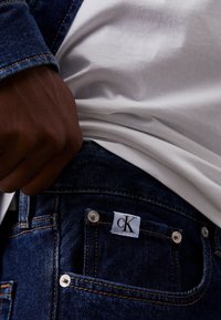 Tmavě modré džínové kalhoty s oranžovým šitím, na kapse s bílou etiketu "cK", stříbrnými nýty a přiléhavým střihem.