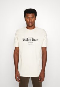 Cremefarbene Baumwoll-T-Shirt mit kurzen Ärmeln, das ein dunkles Druckdesign zeigt, auf dem "CLUB Broken Heart" in stilisierten Schriftarten steht.