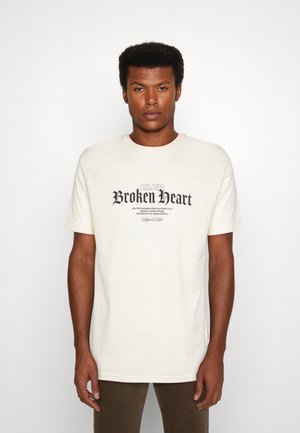 Vaaleancreme värinen puuvillapaita, jossa on lyhyet hihat ja tummaprintattu design, jossa lukee "CLUB Broken Heart" tyylitellyin kirjaimin.