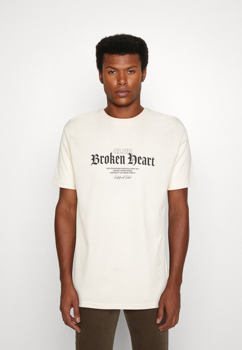 Cremefarbene Baumwoll-T-Shirt mit kurzen Ärmeln, das ein dunkles Druckdesign zeigt, auf dem "CLUB Broken Heart" in stilisierten Schriftarten steht.