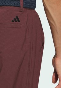 Vinröda byxor med ett strukturerat mönster, med två fickor och en Adidas-logotyp nära vänster ficka. En vävd marinblåt bälte är synlig.
