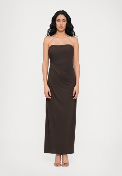 Robe maxi marron sans bretelles avec un corsage ajusté, tissu drapé à la taille et une texture lisse. Portée avec des sandales à lanières de couleur claire.