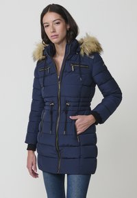 Giacca puffer blu navy con cappuccio in finto pelo, tasche con zip, vita regolabile con cordino e dettagli in oro. Design trapuntato testurizzato.