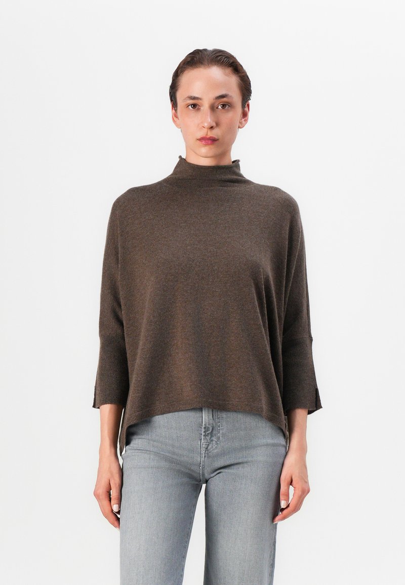 Brauner Strickpullover mit hohem Kragen, Dreiviertelärmeln und seitlichen Schlitzen, kombiniert mit hellgrauen, enganliegenden Jeans. Glatte Textur.