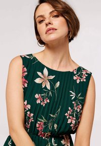 Grünes Blumenkleid mit Falten, kurzen Ärmeln und rundem Halsausschnitt. Verziert mit rosa und weißen Blumen sowie Blättern für visuelle Akzente.