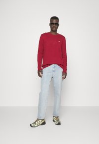 Maglione rosso a maglia, jeans azzurri chiari e sneaker verde camouflage, con un design slim fit. Occhiali da sole completano l'outfit.