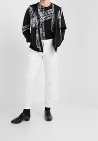 Czarna bluza na zamek z białym napisem, zestawiona z czarną koszulą z grafiką, białymi spodniami i wypastowanymi czarnymi butami. Gładkie tekstury materiałów.