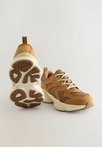 Brązowe sportowe buty na podeście z siateczki i zamszu, kremowa podeszwa środkowa, teksturowana podeszwa z gumy oraz wzmocniony język z tyłu. Widoczne sznurowadła i przeszycia.