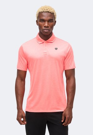 Jeune homme aux cheveux blonds courts portant un polo corail à manches courtes et un pantalon noir, debout devant un fond clair.