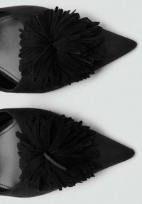 Zapatos negros de punta, hechos de ante, con una gran flor de fieltro negro texturizado como adorno en el empeine. Diseño simple y elegante.