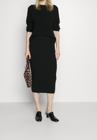 Svart ribbad tröja och figurnära midi-kjol. Modellen håller en väska med utskärning i mönster och bär svarta mjuka skor med låg klack. Enkelt och slankt design.