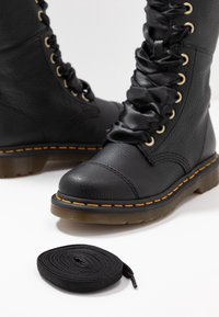 Bottes de combat en cuir noir avec larges lacets en satin et coutures jaunes, accompagnées de lacets ronds noirs supplémentaires sur une surface blanche.